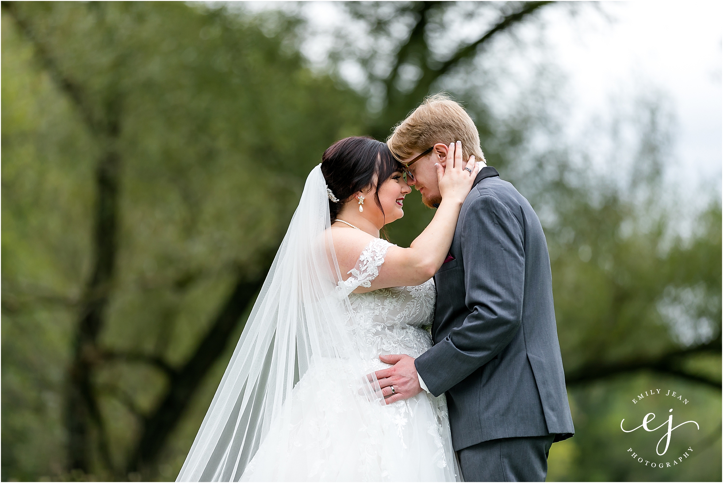 bride holding grooms head and face close to hers in front of large green tree at