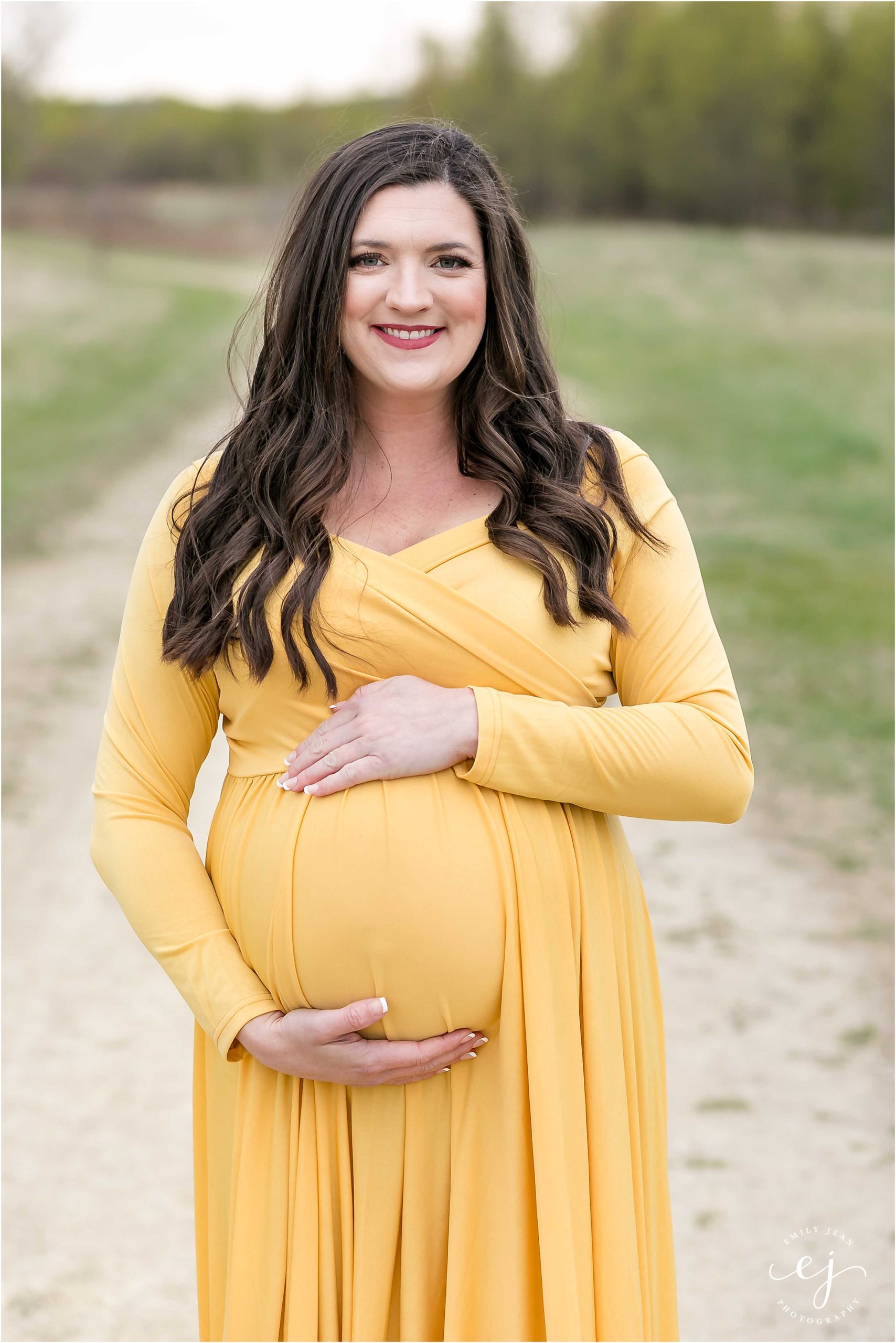 Pregnant woman wearing yellow dress with brown hair smiling at the camera holding belly
