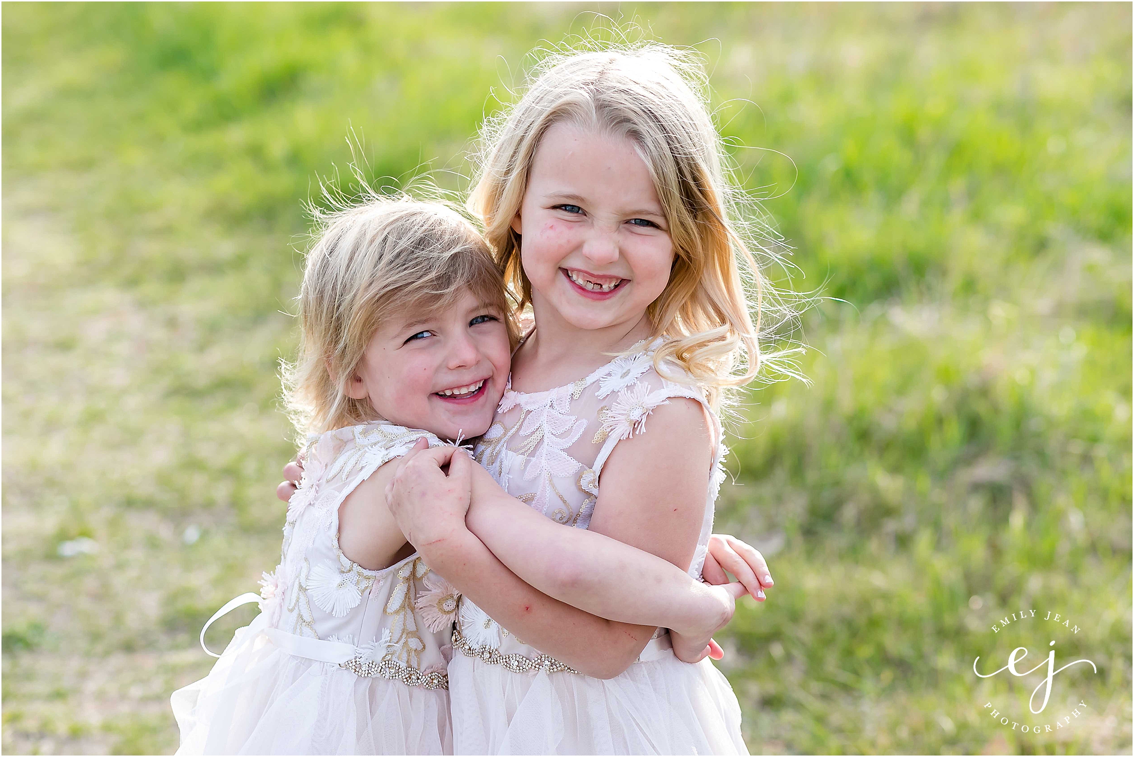 Two cute little blonde sisters hugging each other and smiling at the camera during family photos in the spring time