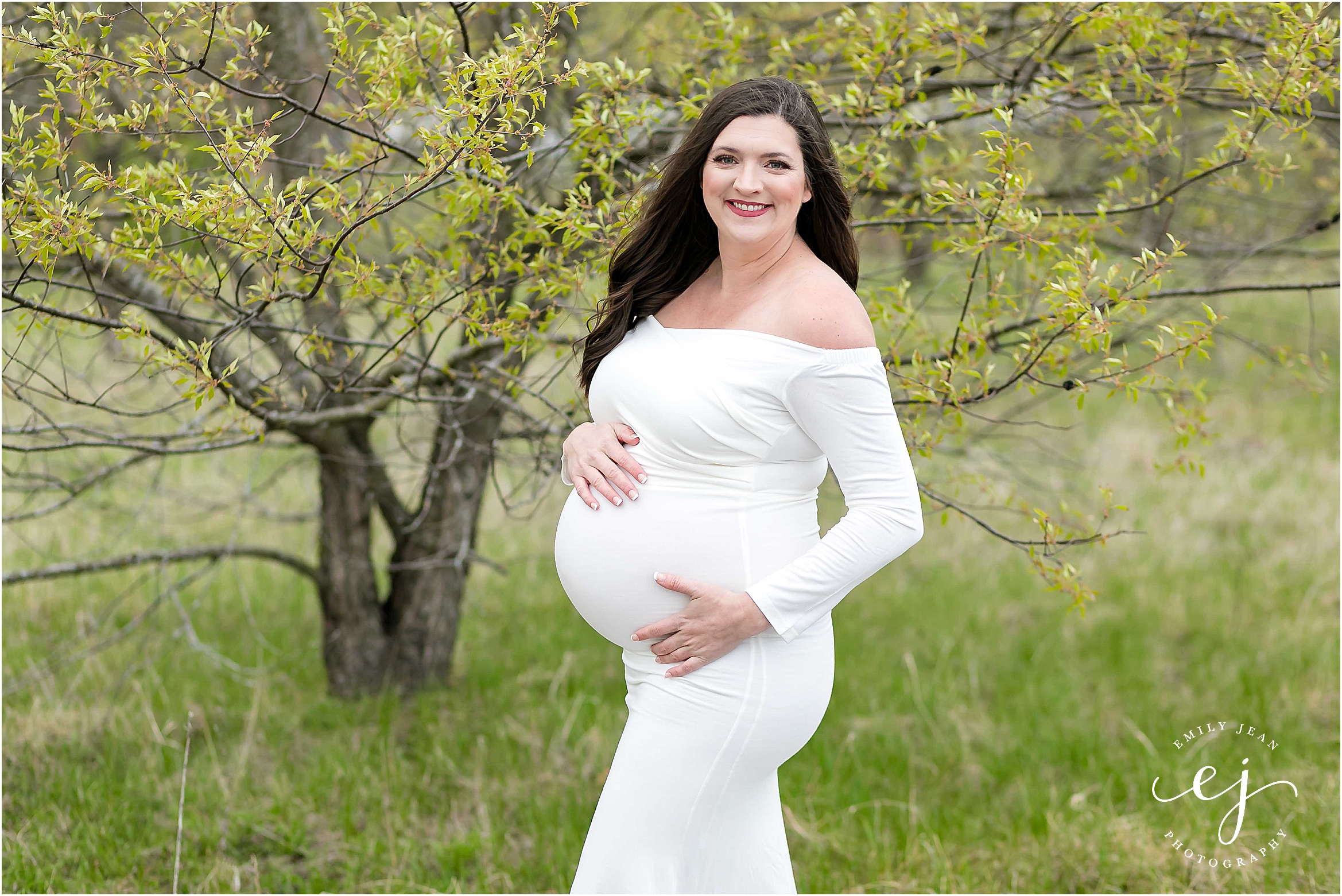 Spring maternity photo woman wearing white dress standing near a green tree